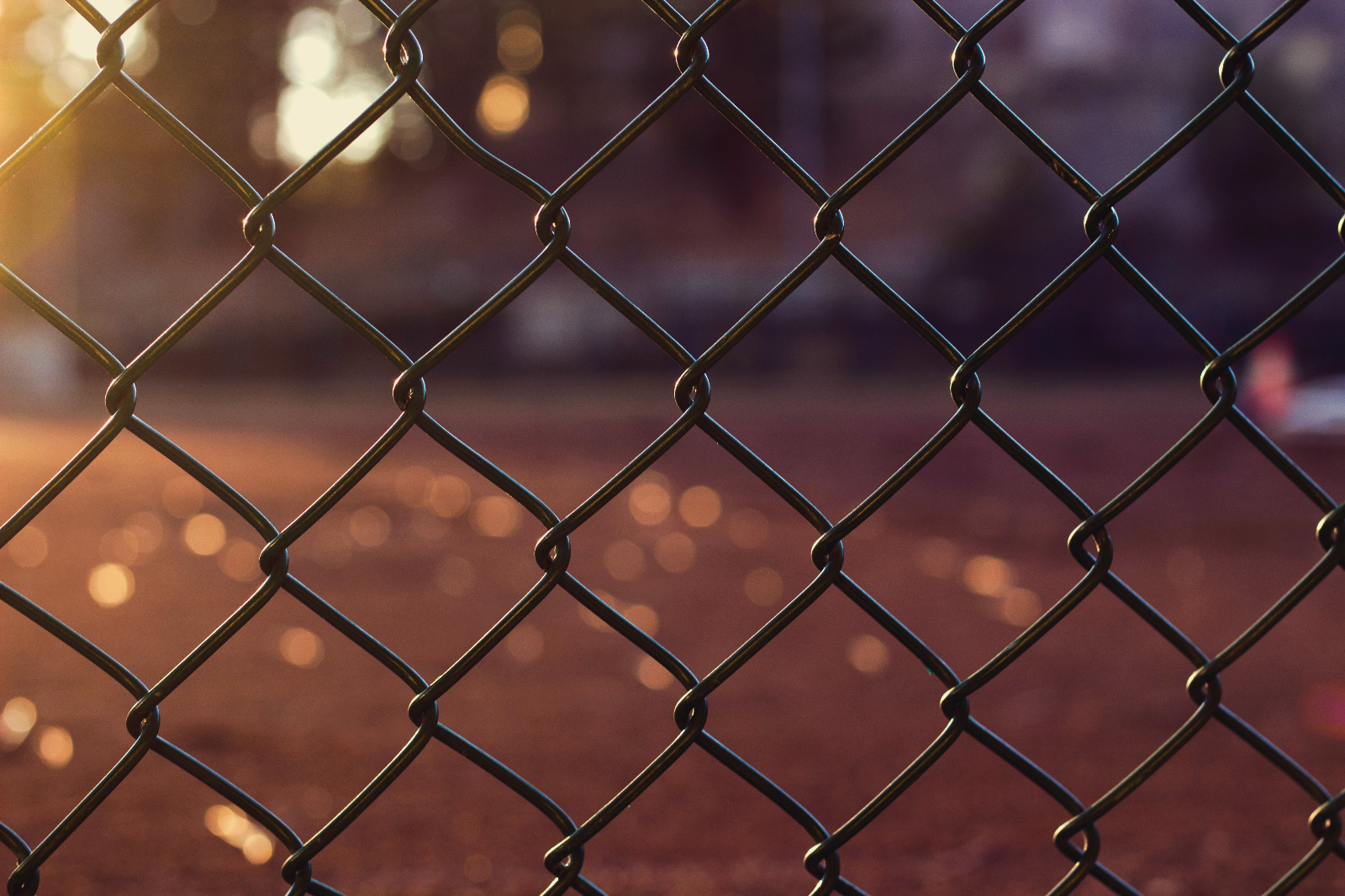 A close-up view of a chain-link fence with sunlight shining on a blurred brown ground in the background.