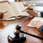 Two people discuss legal documents at a desk with a judge's gavel and scales of justice visible.