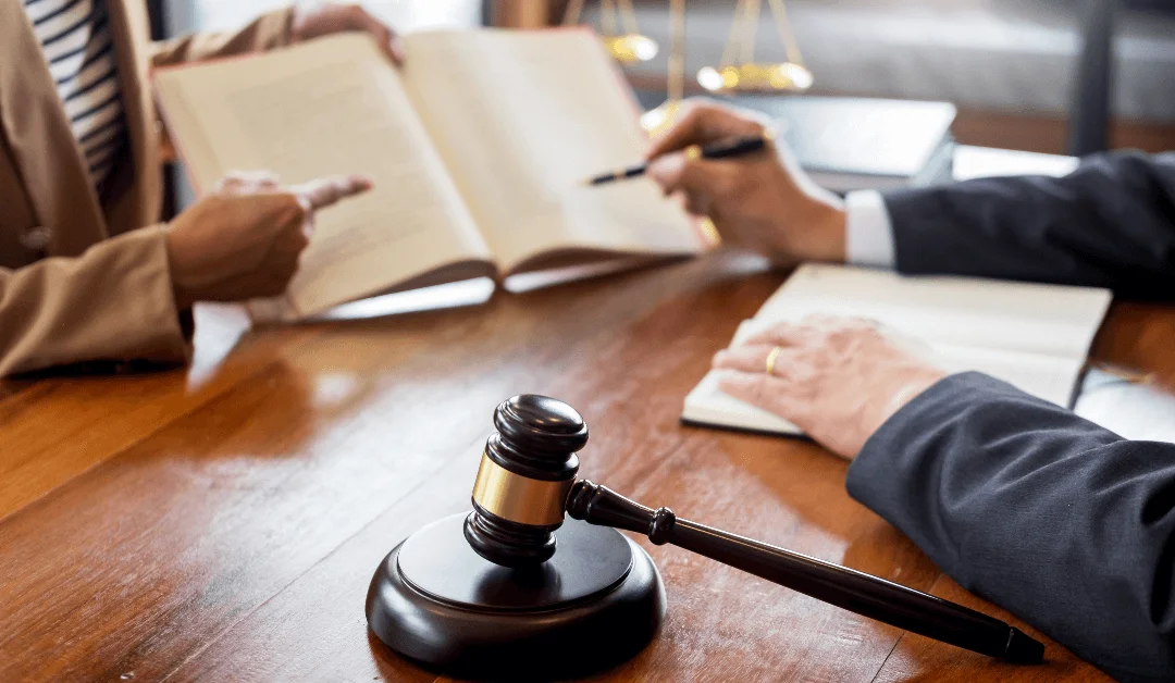 Two people discuss legal documents at a desk with a judge's gavel and scales of justice visible.